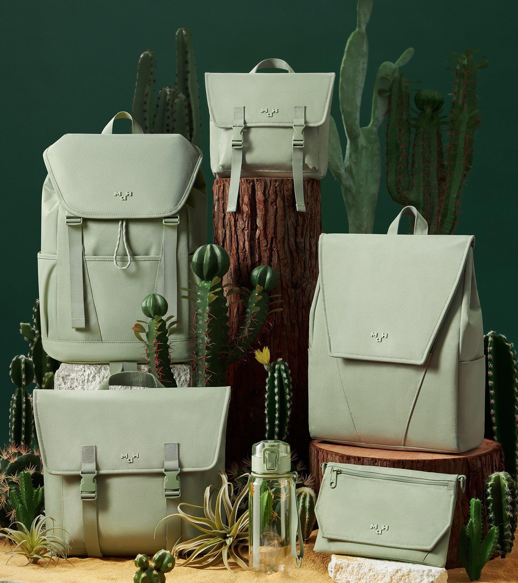 Sage green backpacks, bags, and accessories displayed with cacti and wood stumps for a desert-inspired lifestyle look