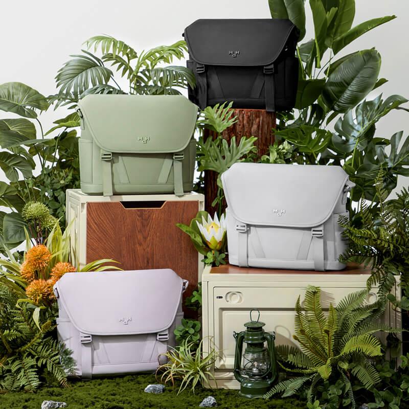 Stylish messenger bags in black, sage, white, and lavender displayed among lush green plants and vintage furniture