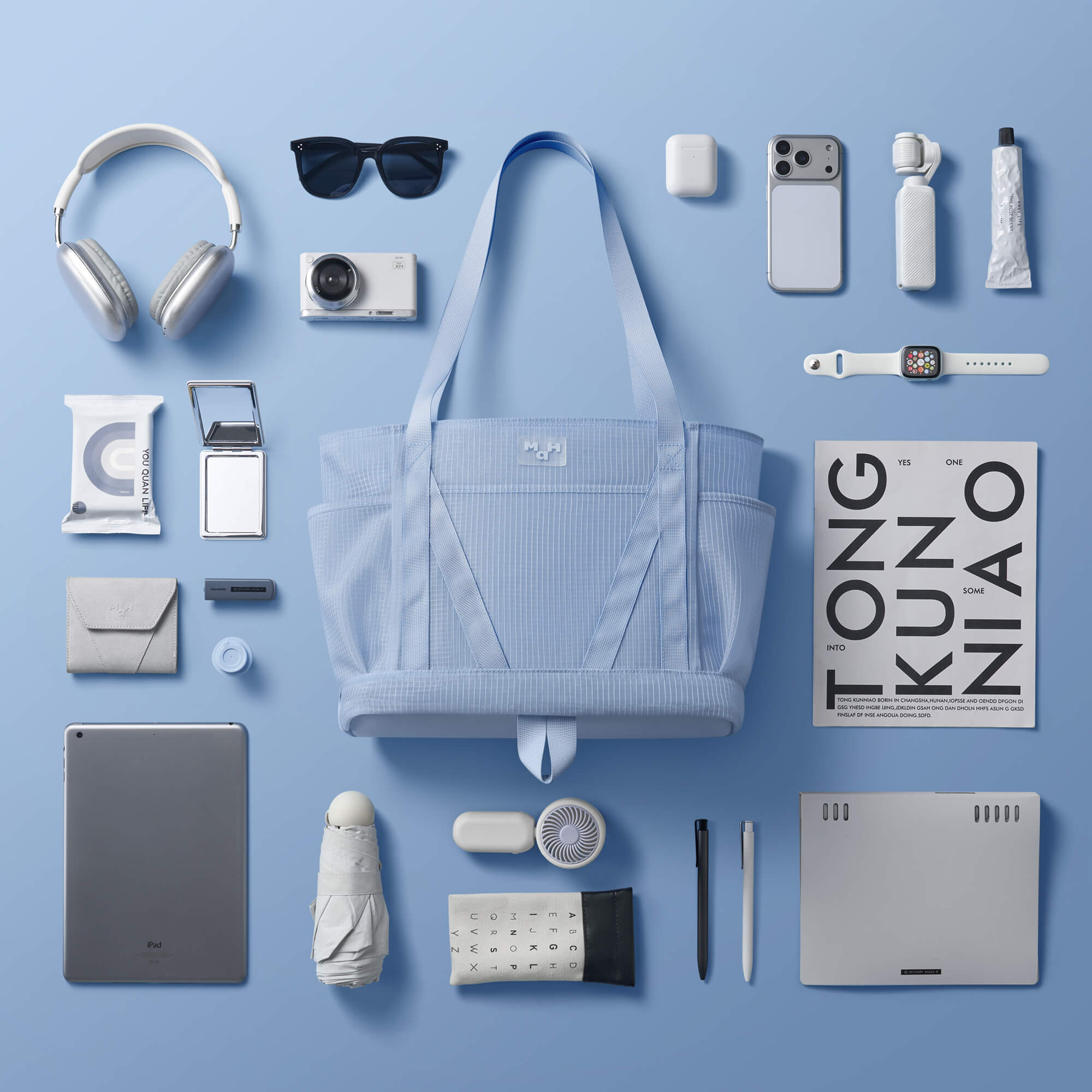 Light blue striped tote bag with headphones, sunglasses, phone, tablet, and travel essentials on blue background