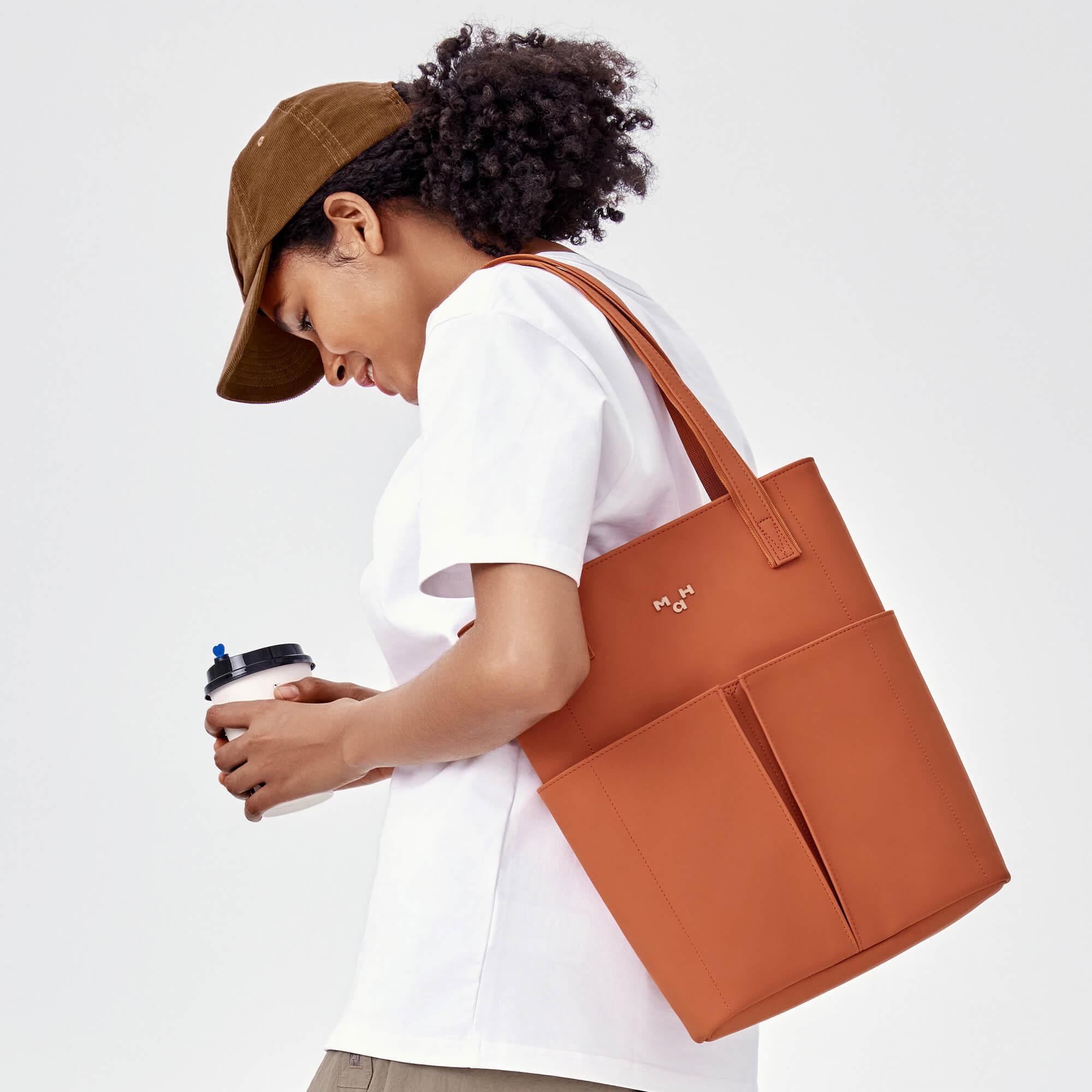 Woman carrying stylish terracotta tote bag with front pockets, holding coffee cup, wearing cap and white tee