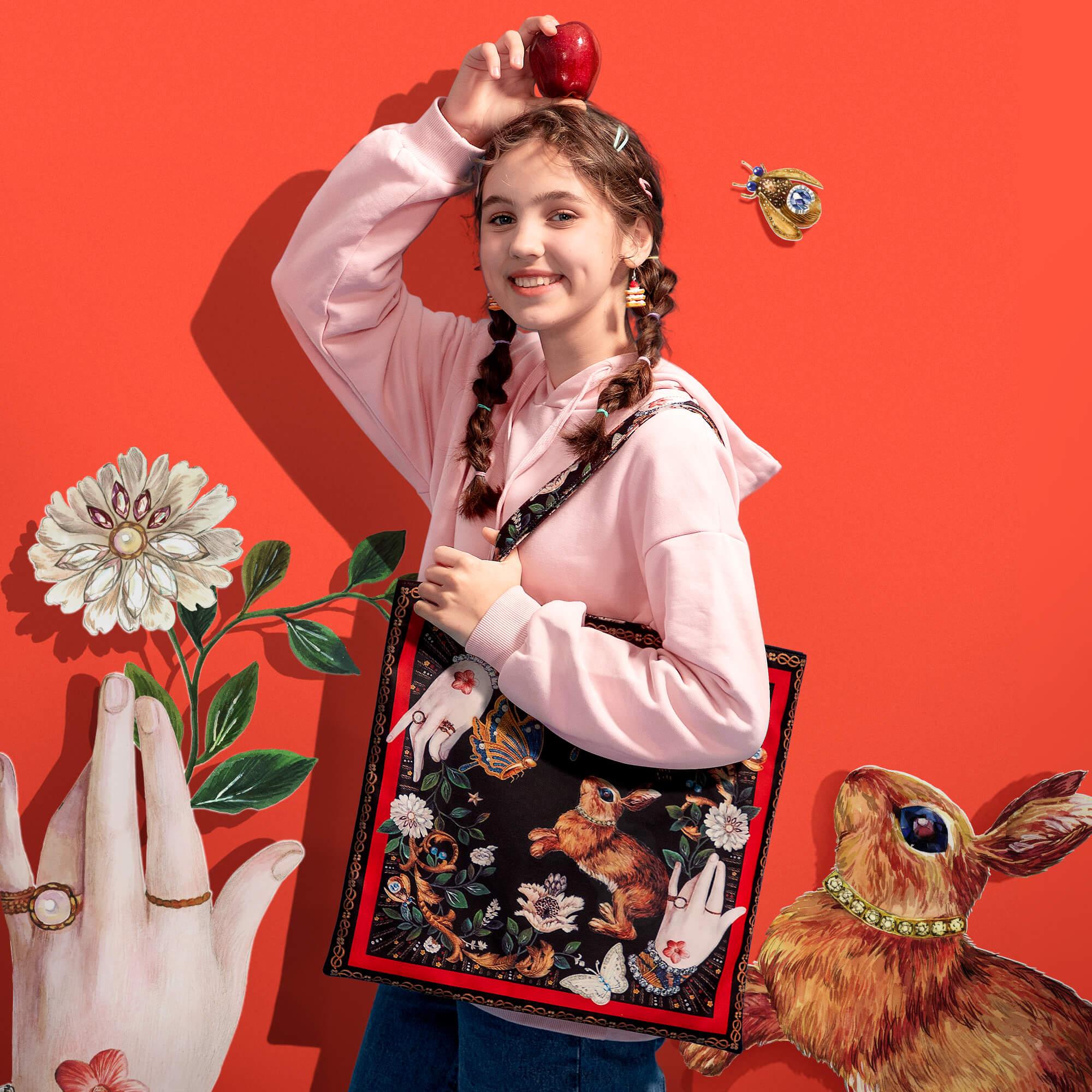 Girl holding apple with floral and rabbit print tote bag, styled with braids and pink hoodie