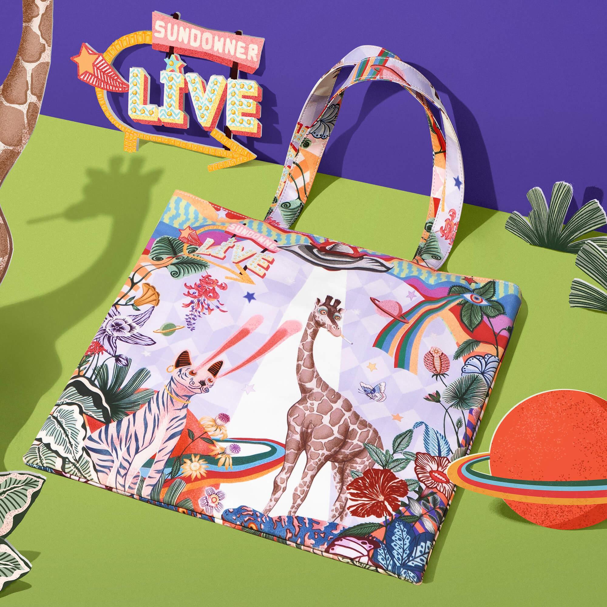 Colorful tote bag with giraffe, cat, rainbow, and floral print, styled with 'Sundowner Live' sign on purple and green background
