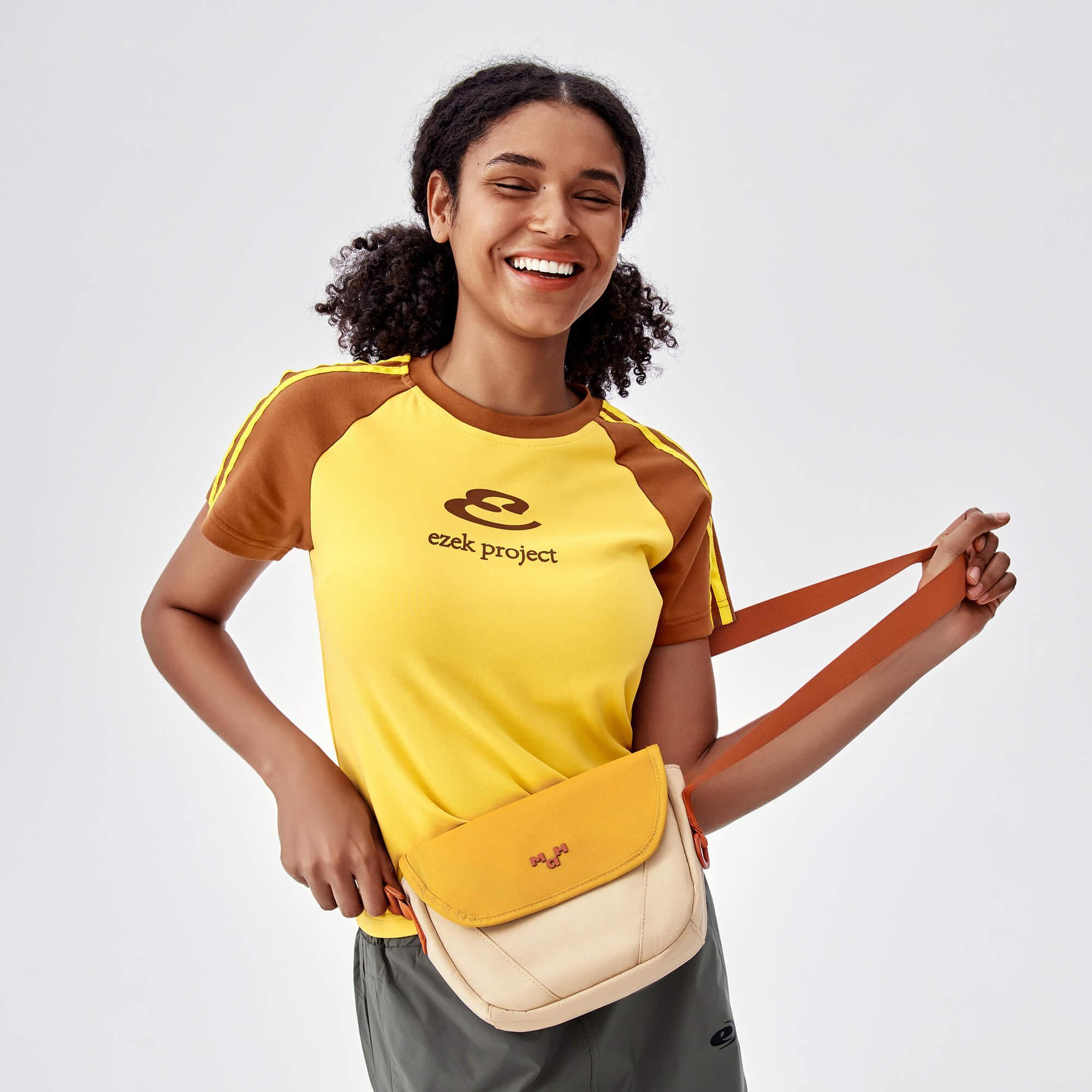 Woman smiling with yellow and beige crossbody bag, wearing ezek project t-shirt, lifestyle accessory