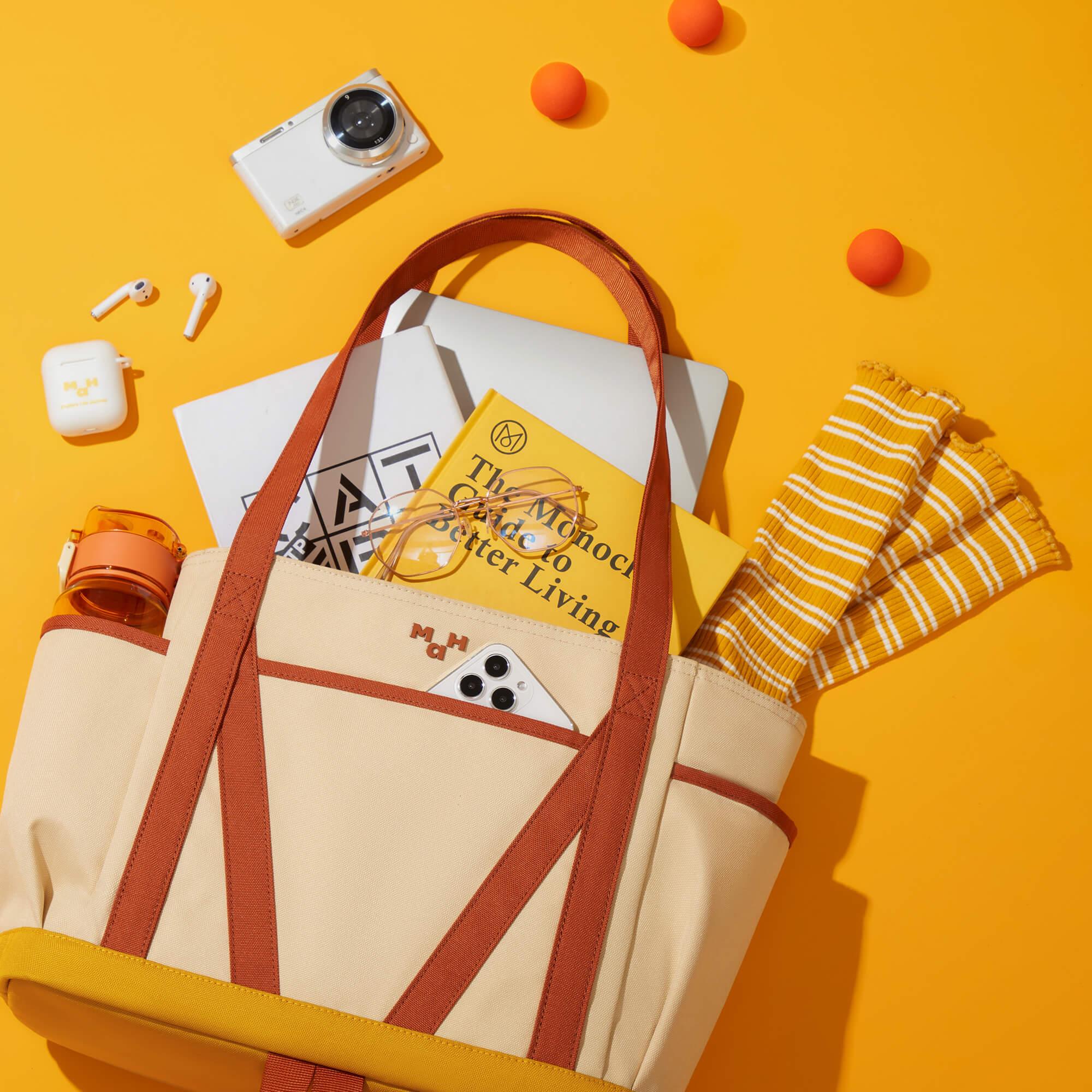 Beige and orange tote bag with laptop, book, phone, and accessories on yellow background