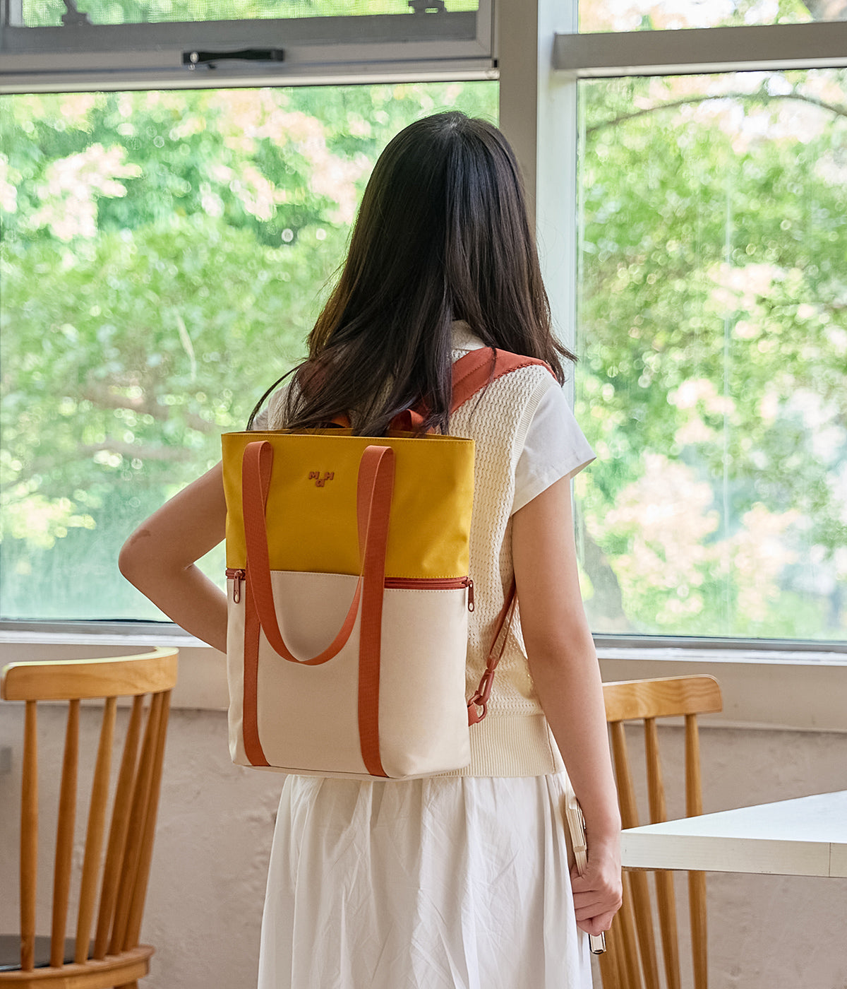 Woman wearing a yellow and cream two-tone school backpack while standing by a window.