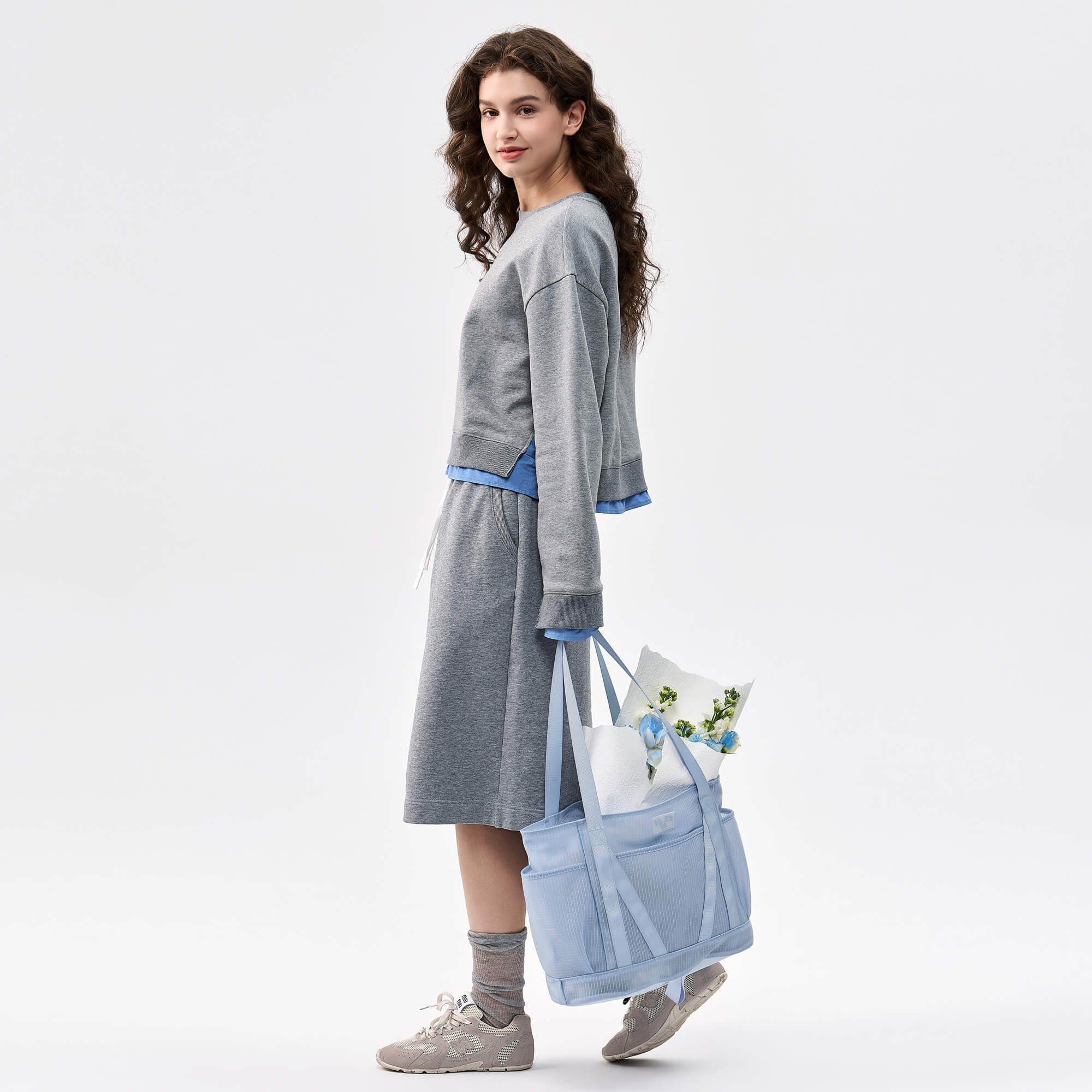 Woman carrying light blue mesh tote bag with flowers, styled with gray sweatshirt dress and sneakers