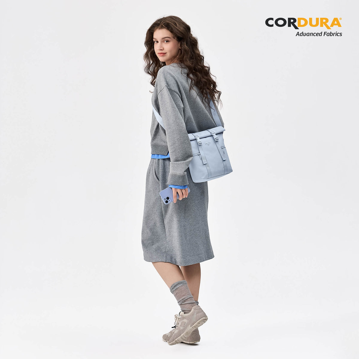 Woman wearing light blue Cordura crossbody bag with gray outfit, holding phone, stylish everyday accessory