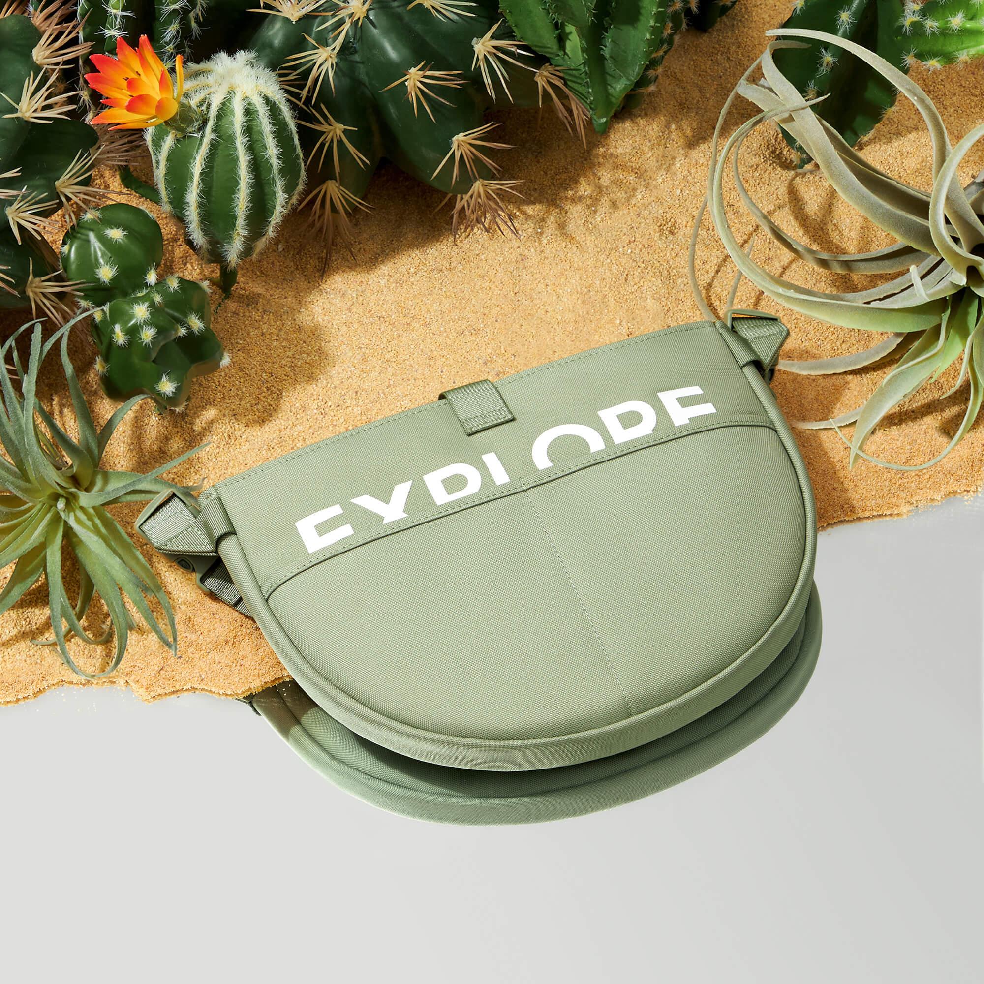 Olive green crossbody bag with white text on sandy desert background with cacti and succulents