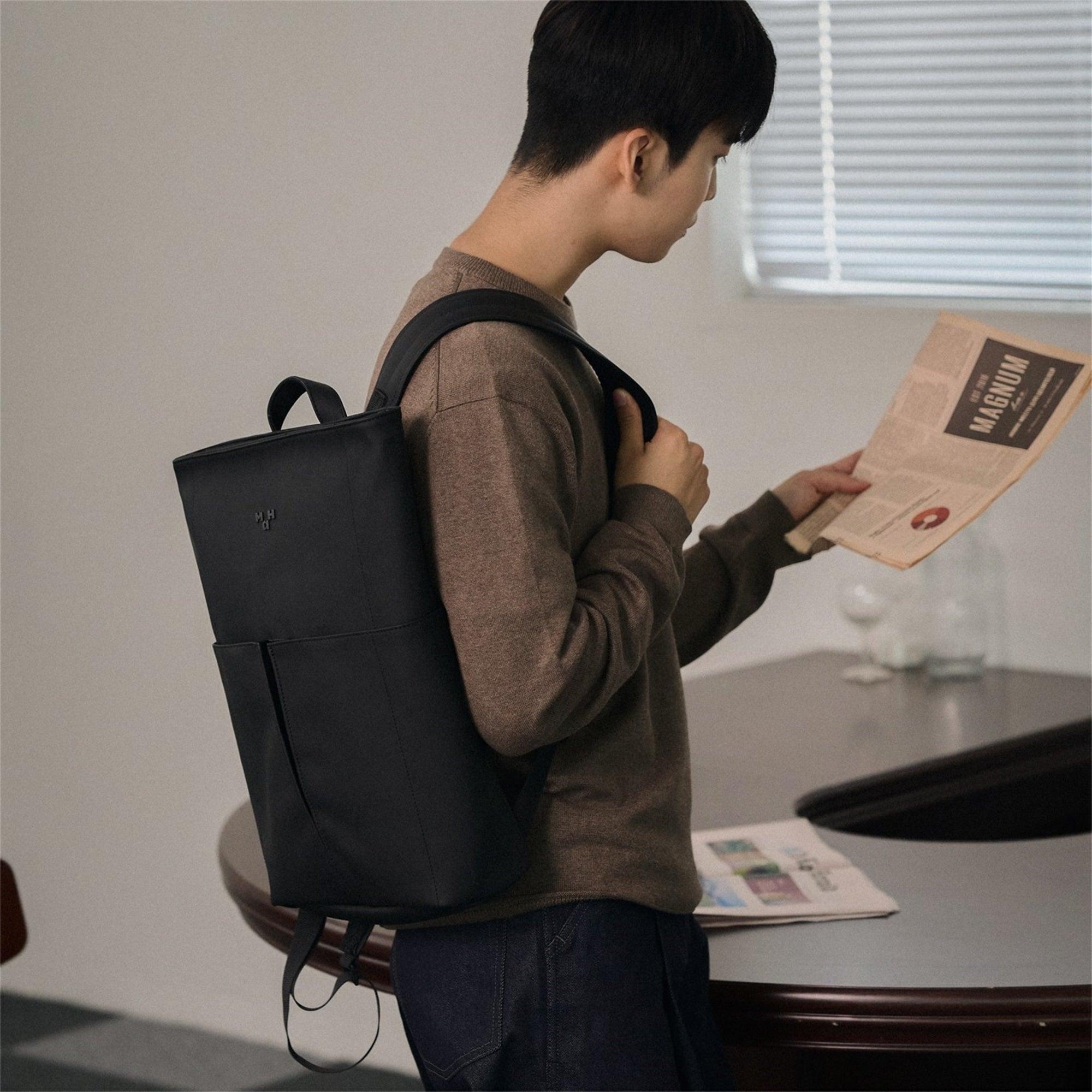 Man wearing sleek black minimalist backpack, reading newspaper in modern indoor setting