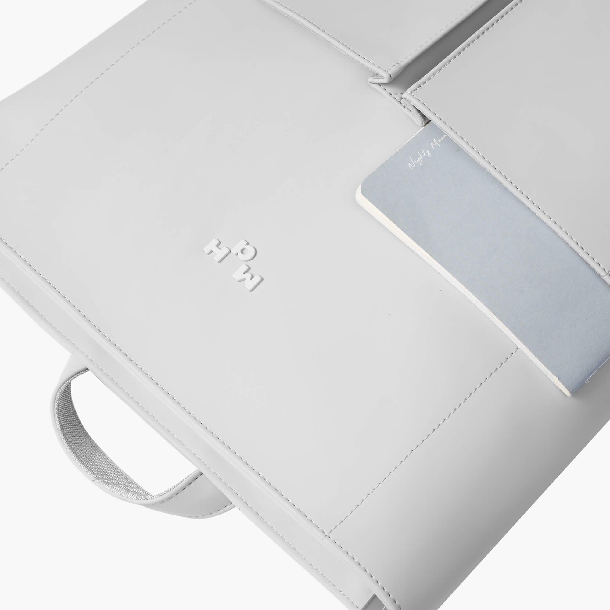 Light gray minimalist backpack with embossed logo and front pocket holding a notebook, ideal for daily use or travel.