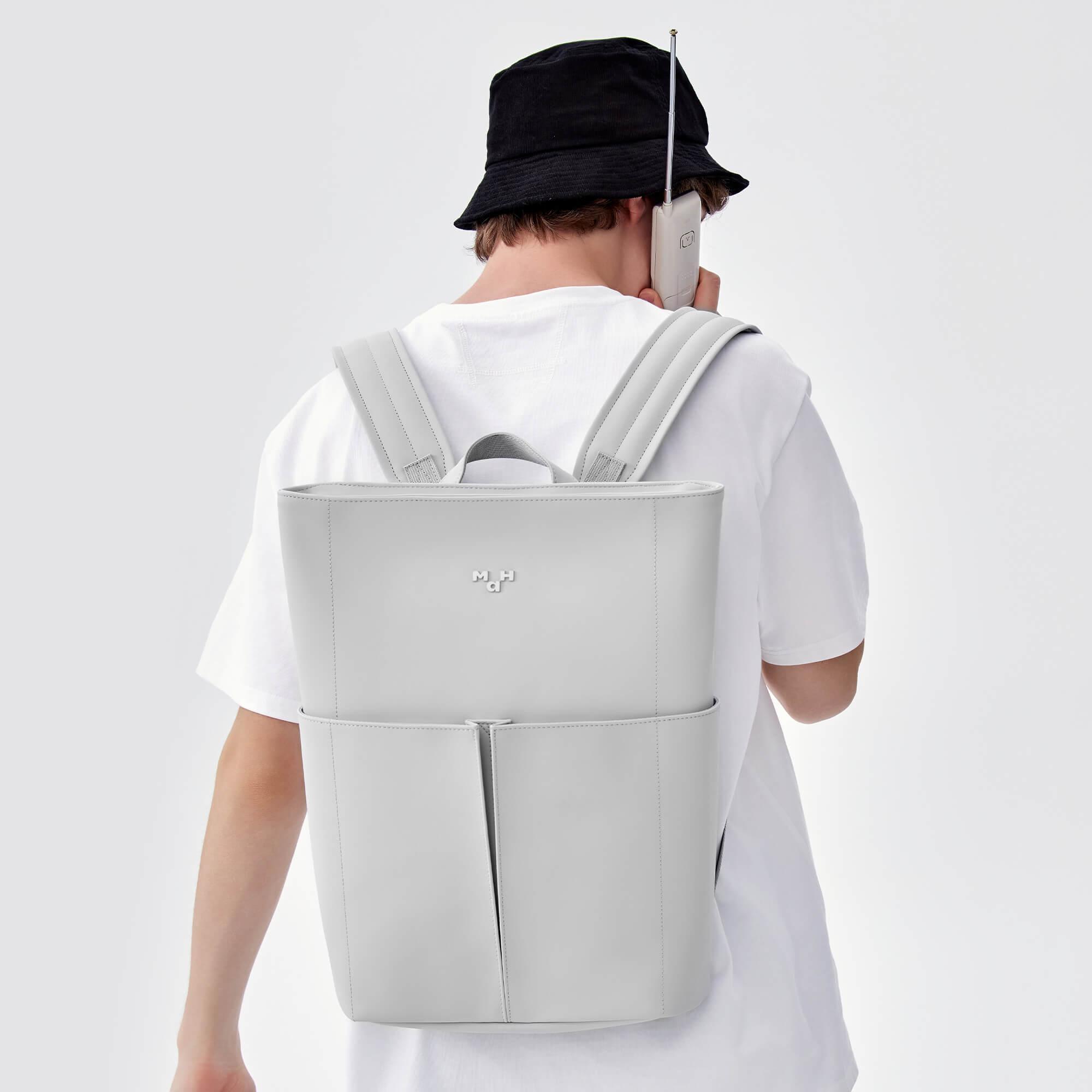 Light gray minimalist backpack with front pockets, worn by person in white tee and black hat