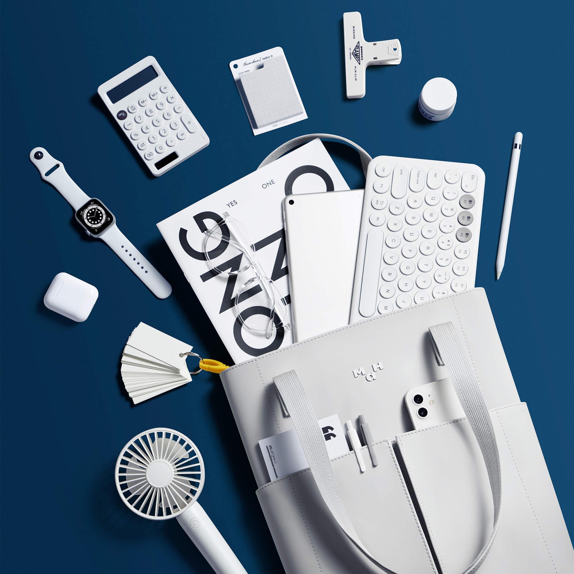 Minimalist white desk setup with laptop, keyboard, calculator, smartwatch, and sleek tote bag on blue background