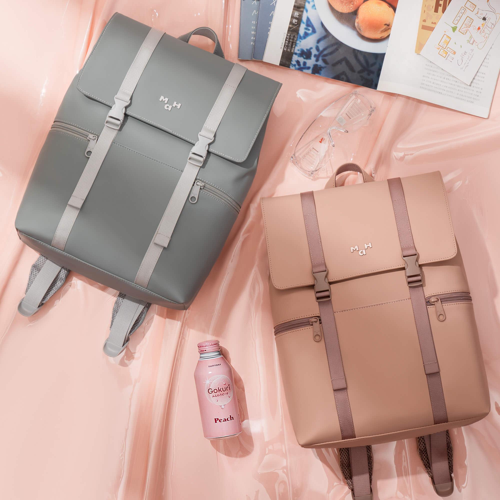 Stylish gray and blush pink backpacks with buckles, perfect for daily use or school, styled with magazines and a peach drink.