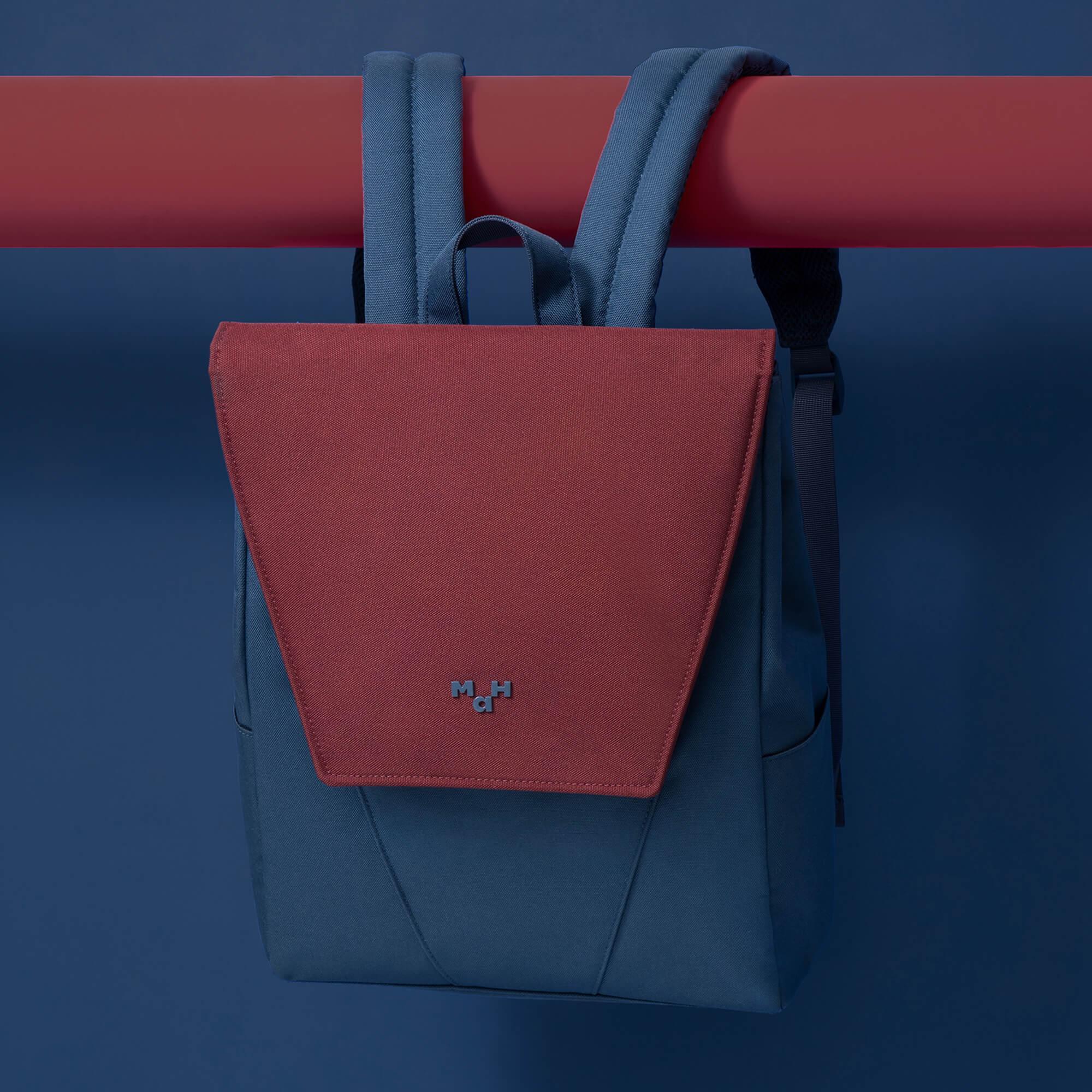Red and blue backpack with M H logo, hanging on red bar against dark blue background