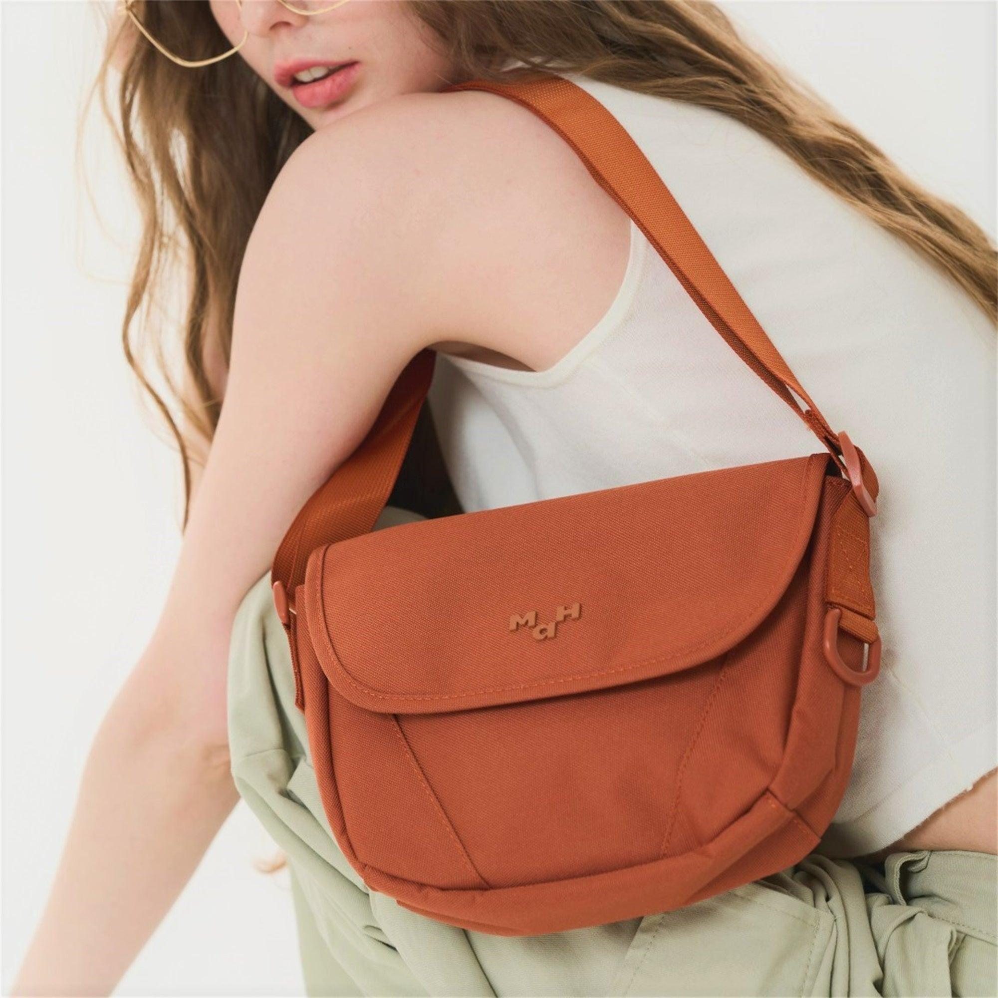 Rust-colored crossbody bag with adjustable strap, worn by woman in white top, featuring minimalist design and subtle logo.