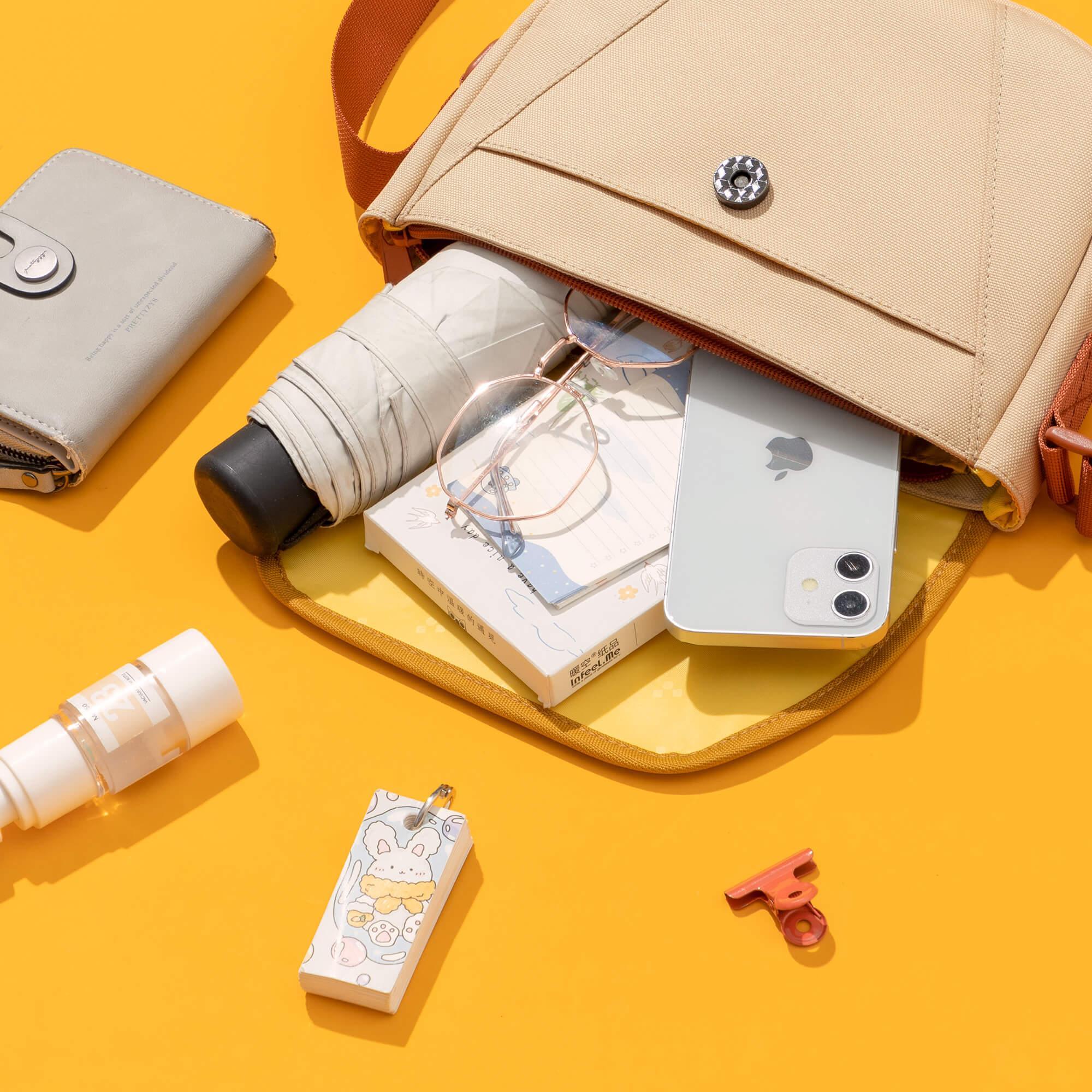 Beige crossbody bag with phone, notebook, umbrella, and accessories on yellow background
