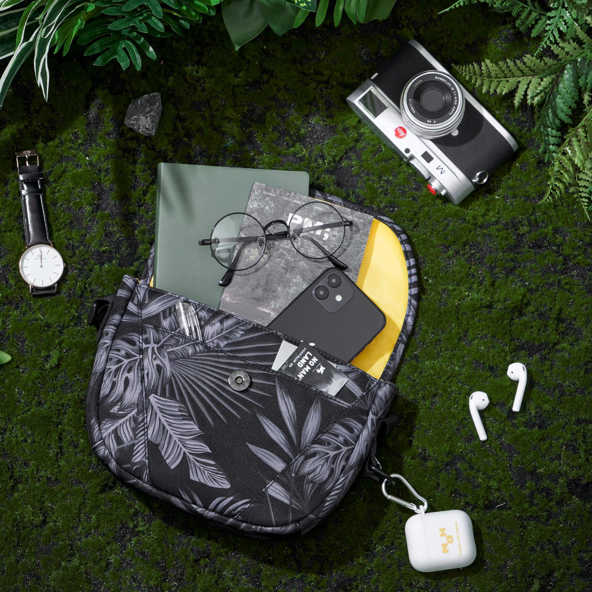 Tropical print sling bag with phone, glasses, notebook, camera, watch, and wireless earbuds on mossy ground
