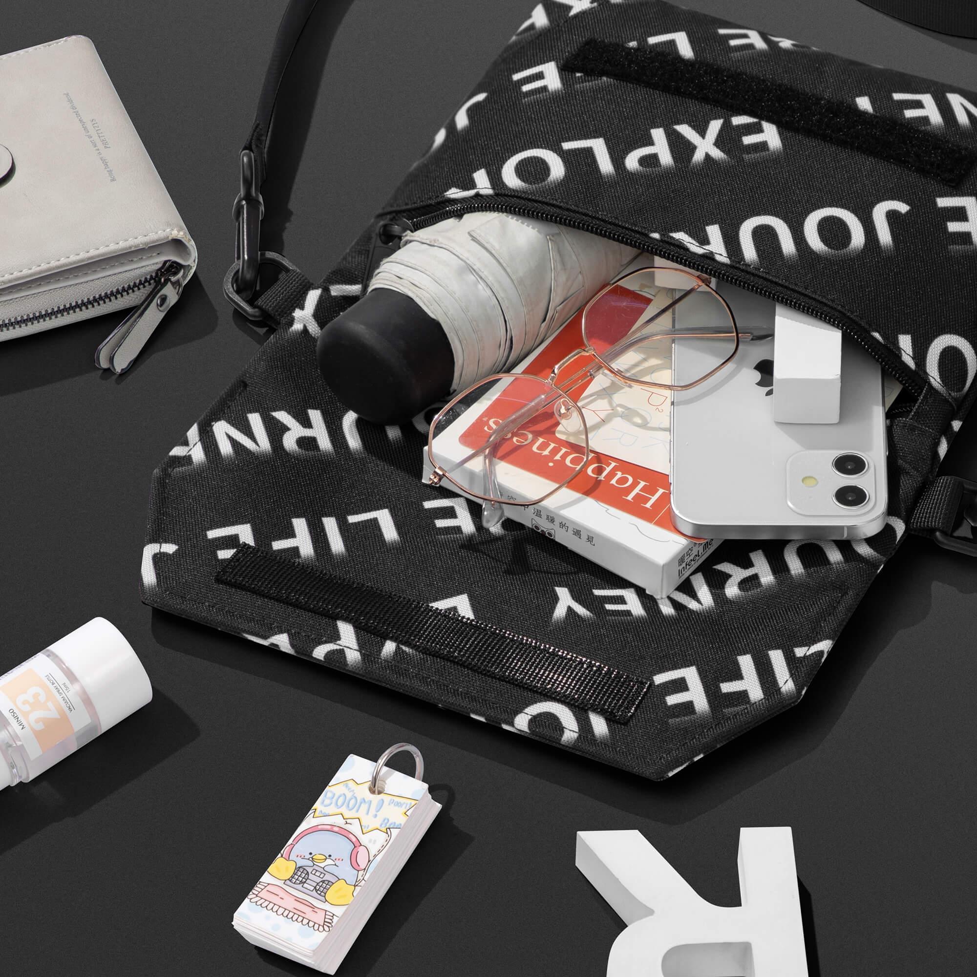 Black crossbody bag with white text print, holding phone, book, glasses, and umbrella, styled with wallet and accessories