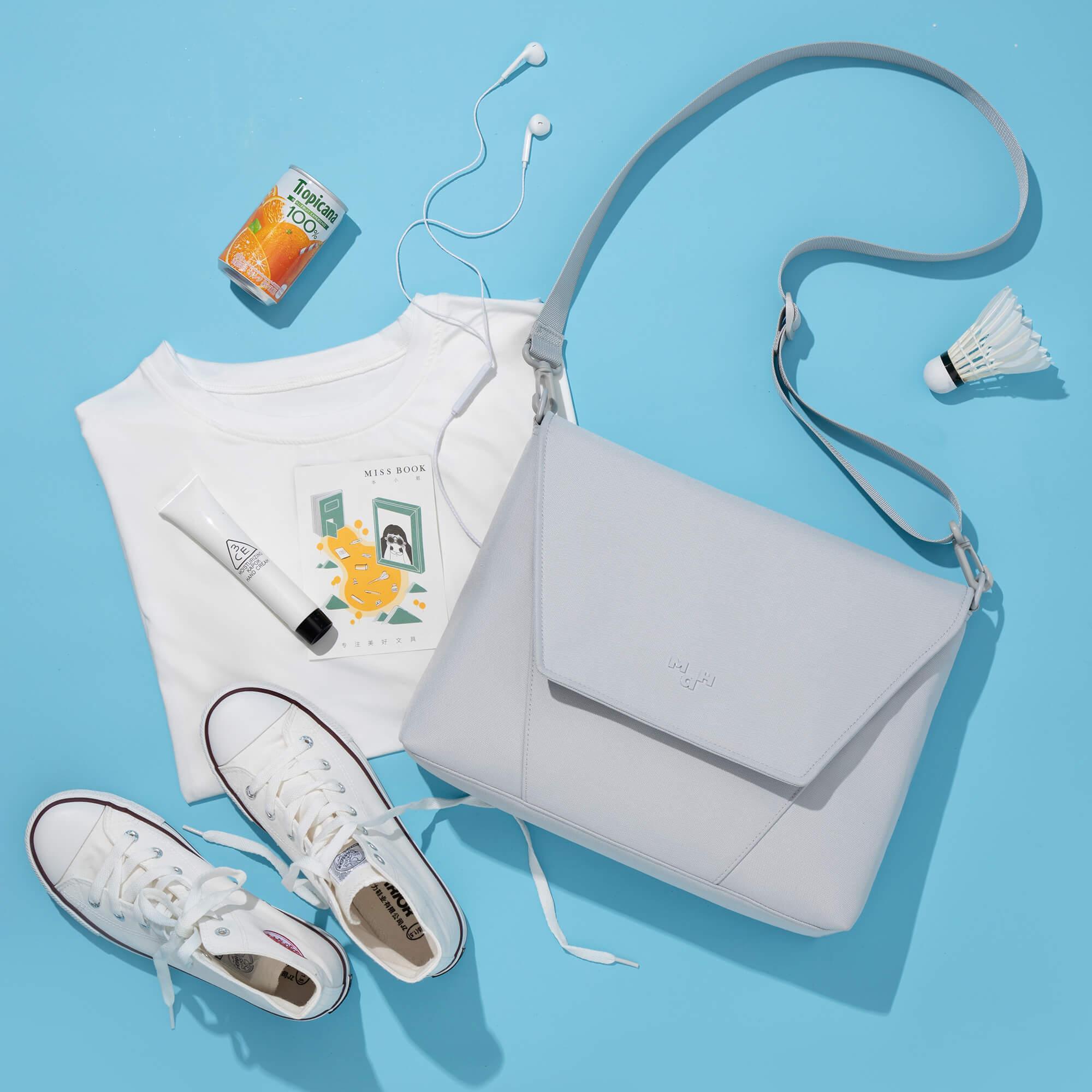 Light gray crossbody bag with white sneakers, T-shirt, earphones, and orange juice on blue background