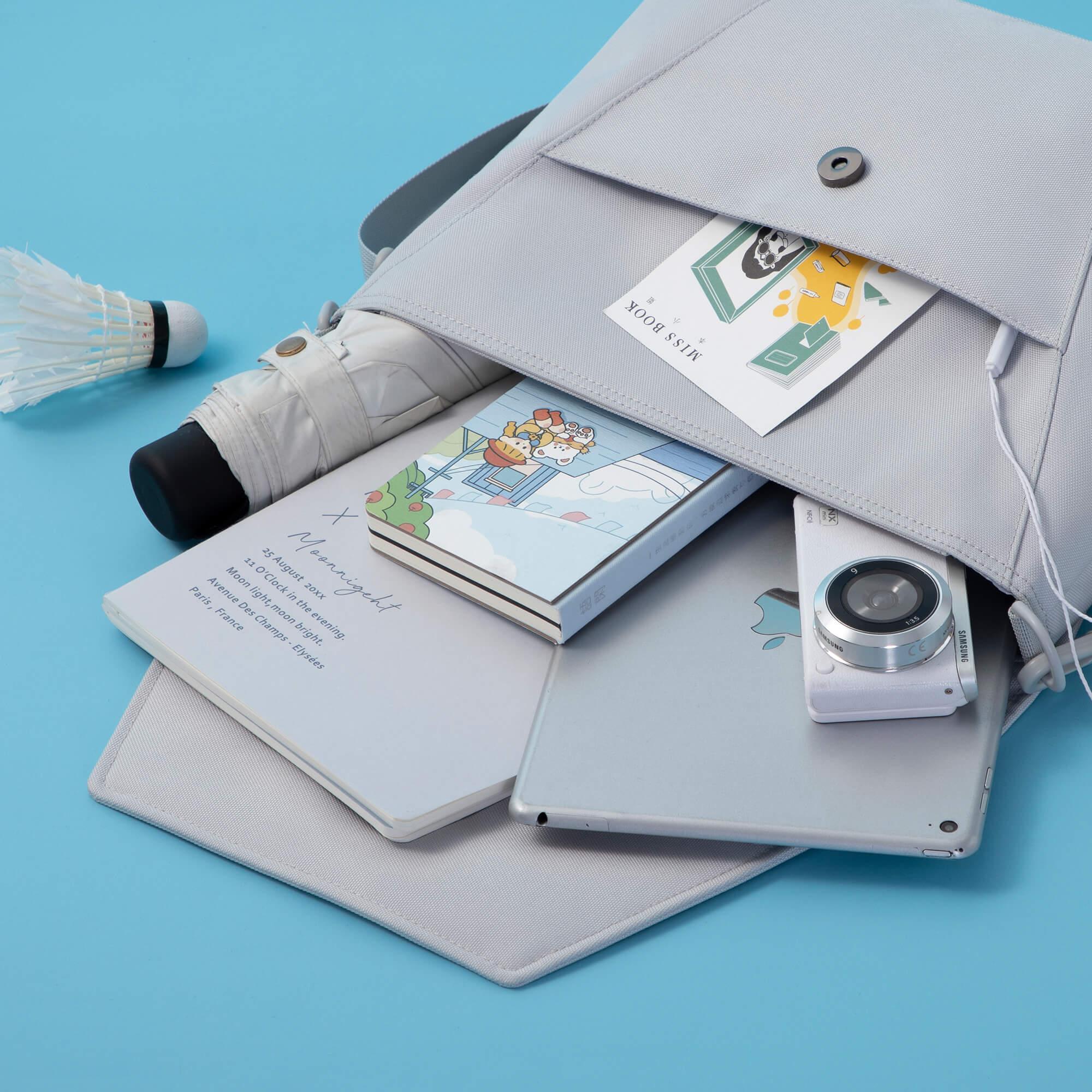 Light gray canvas bag holding notebook, camera, tablet, and umbrella on blue background