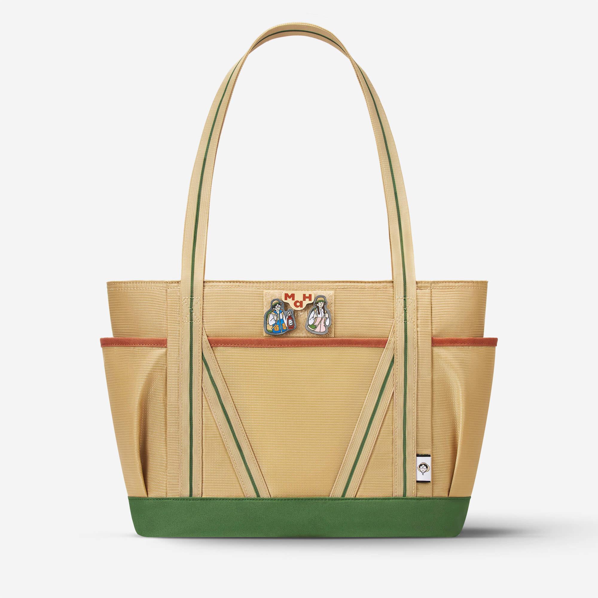 Beige canvas tote bag with green trim, front pockets, and character pins, ideal for everyday use or travel.