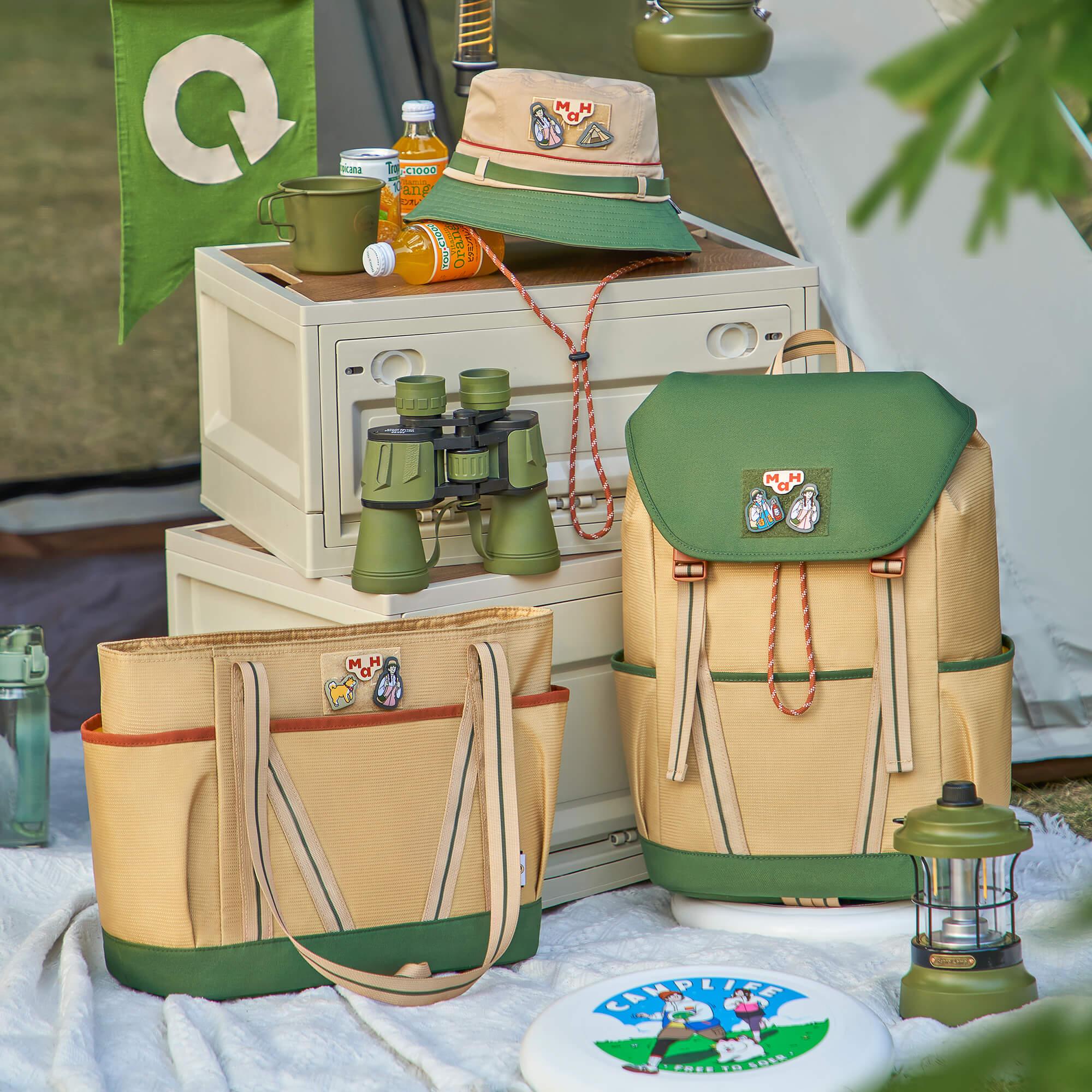 Green and beige camping backpack and tote bag with patches, binoculars, hat, and lantern at campsite