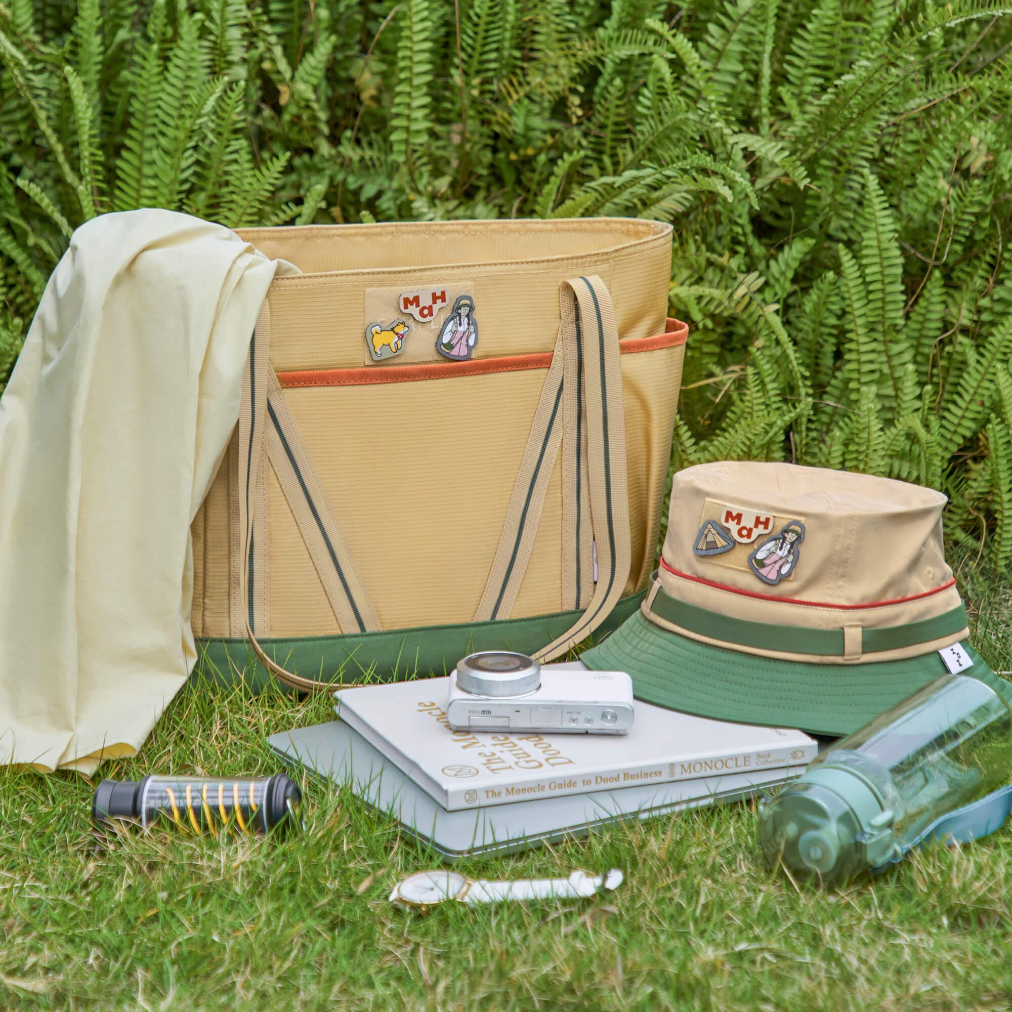 Beige tote bag with patches, bucket hat, books, and camera on grass for outdoor lifestyle