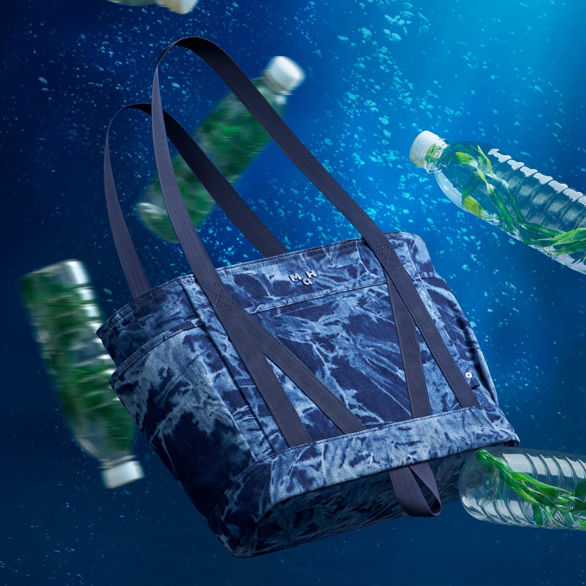 Blue tie-dye tote bag with black straps floating underwater near plastic bottles, eco-friendly design