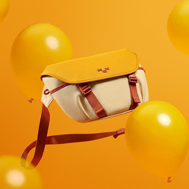 Yellow and cream crossbody bag with red straps, floating among yellow balloons on orange background